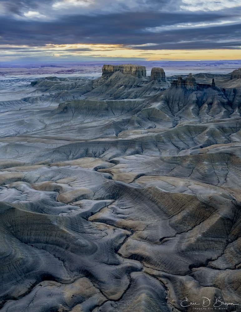 Lunar Landscape: Twilight at Moon Overlook -  
The otherworldly badlands near Hanksville, Utah stretch toward the horizon as dusk settles over the terrain, casting the eroded channels and mesas in cool blue tones while a sliver of golden light breaks through the cloud cover, illuminating the distant formations.